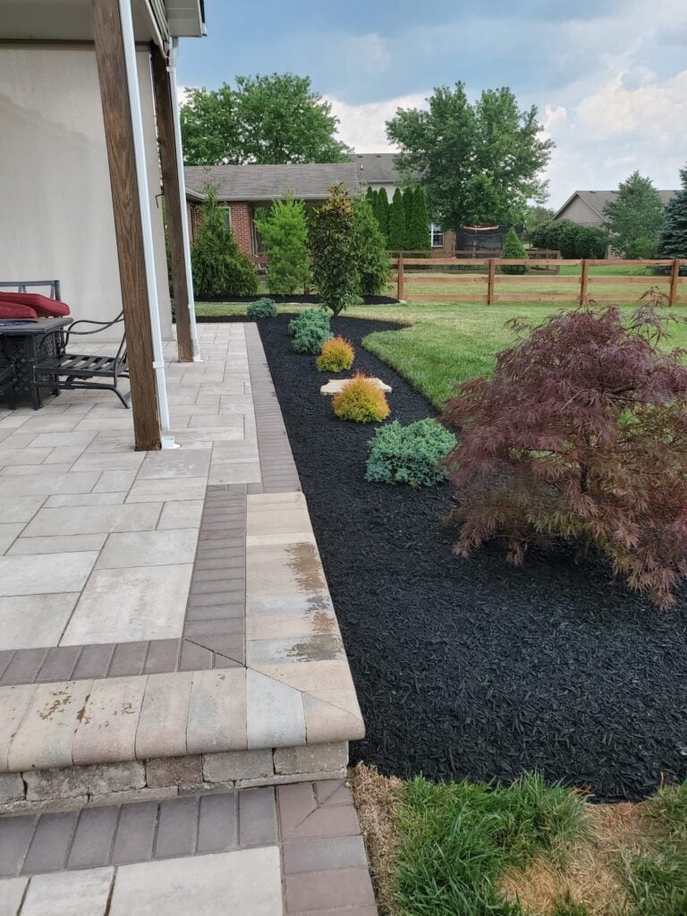 Landscaped backyard with a paved patio, mulched planting beds, shrubs, lawn, and a wooden fence