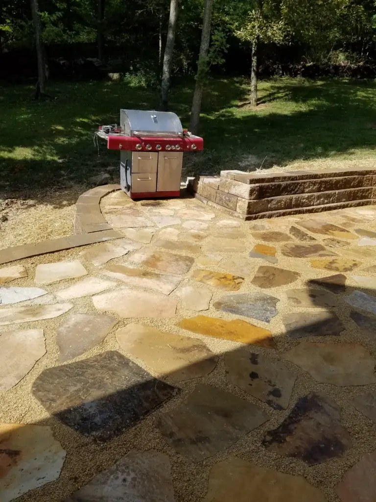 Stone patio with a grill beside a low retaining wall and lawn
