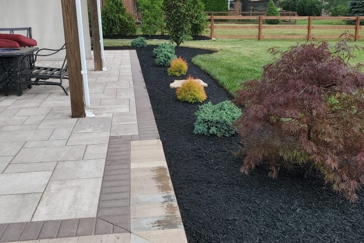 Landscaped backyard with a paved patio, mulched planting beds, shrubs, lawn, and a wooden fence