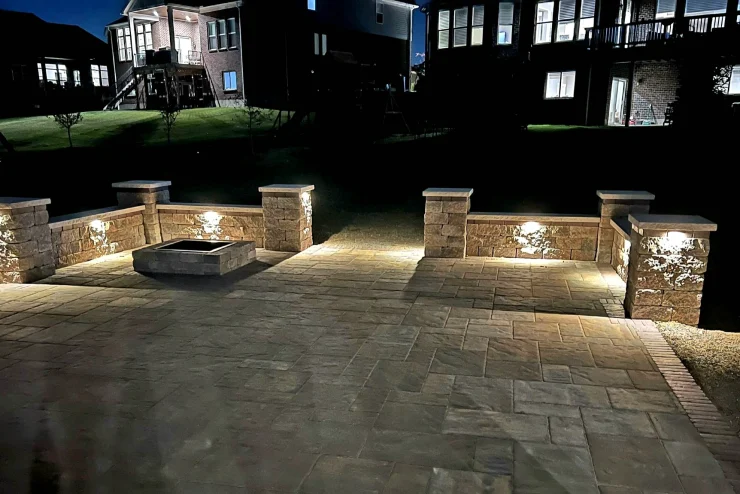 Illuminated stone patio with low seating walls and a square fire pit at night
