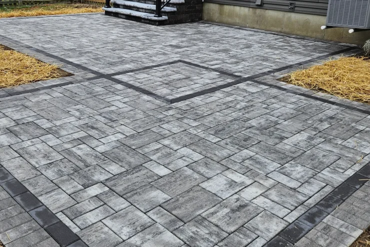 Gray interlocking paver patio with dark edging beside a house and landscaped beds