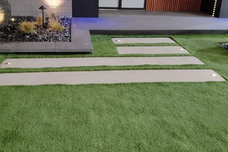 Landscaped yard with artificial grass, concrete stepping paths, a rock garden, and outdoor lighting